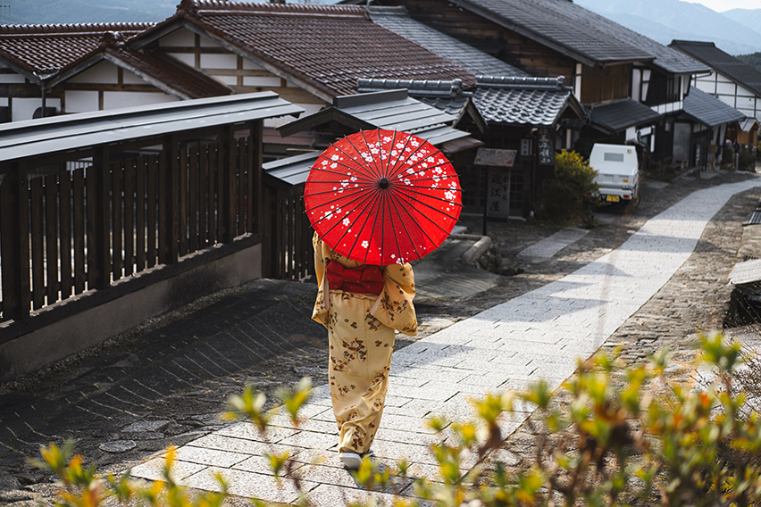 Foto de uma mulher vestida tradicionalmente passeando em uma das ruas tradicionais da cidade de Kyoto enquanto se protege do sol utilizando um guarda-sol tradicional do Japão.