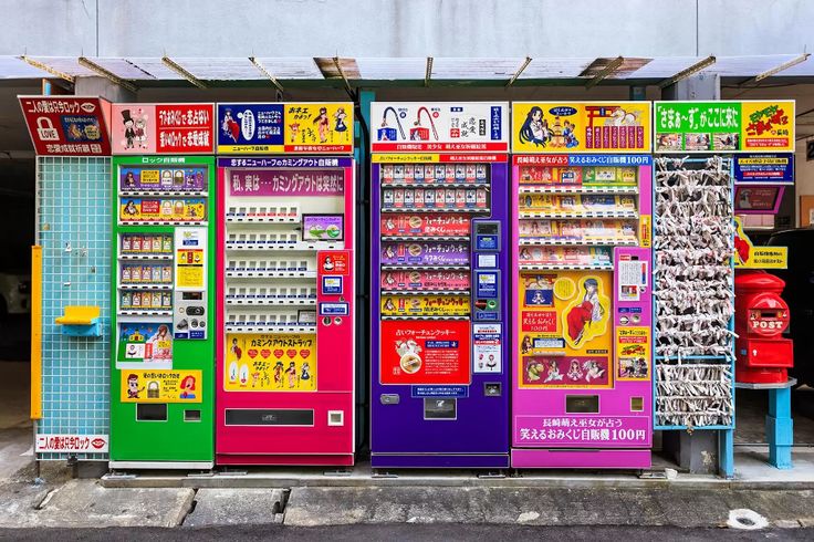 Fila de máquinas de vendas automaticas de várias marcas e tipos de bebidas em uma rua de pequena cidade no Japão. ao fundo podemos ver um carro típico dessas pequenas cidades ou vilarejos.