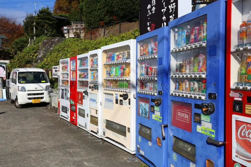 Fila de máquinas de vendas automaticas de várias marcas e tipos de bebidas em uma rua de pequena cidade no Japão. ao fundo podemos ver um carro típico dessas pequenas cidades ou vilarejos.