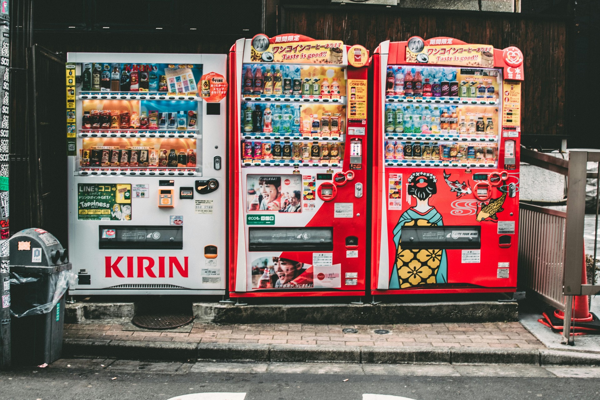 Três máquinas de venda automática em uma calçada, com uma variedade de bebidas. A máquina à esquerda é branca com a marca "KIRIN", enquanto as duas à direita são vermelhas, uma delas com uma ilustração de uma mulher em trajes tradicionais japoneses.